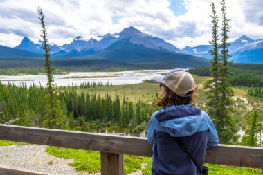 Beyzbol şapkalı ve açık mavi ceketli bir turist, ahşap korkuluklara yaslanmış, kuzey Saskatchewan nehrinin ve Banff Milli Parkı 'ndaki Kanada kayalıklarının panoramik manzarasını izliyor.