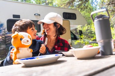 Anne ve oğul Banff Ulusal Parkı 'ndaki karavanın dışındaki piknik masasında kahvaltı ederken birlikte gülüyorlar. Aile bağlarının neşeli bir anını yakalıyorlar.