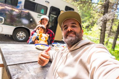 Mutlu bir aile, ormandaki karavanlarının yanındaki ahşap bir masada yemek yerken selfie çekiyor. Banff Ulusal Parkı 'nda tatillerinin tadını çıkarıyorlar. Kanada kayalıkları.