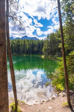 Herbert Gölü 'nün zümrüt yeşili suları bulutlu mavi gökyüzünü ve arka planda kayalık dağlarla çevrili çam ormanlarını yansıtıyor. Banff Ulusal Parkı, Alberta, Kanada