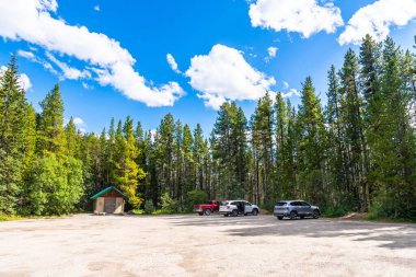 Herbert Gölü otoparkı gür bir çam ormanının altında canlı mavi bir gökyüzü altında kabarık bulutlarla, Banff Ulusal Parkı 'nın güzelliğini keşfetmek için sakin bir zemin sağlıyor.