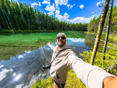 Fotoğrafçı, Herbert Gölü 'nde uzanan kollarıyla selfie çekiyor. Etrafı turkuaz su, yemyeşil orman ve muhteşem bir gökyüzü ile çevrili. Banff Ulusal Parkı' nda, Kanada kayalıklarında, Alberta 'da.