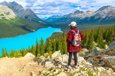 Sırt çantası takan kadın yürüyüşçü, kayalık bir bakış açısında duruyor ve Banff Ulusal Parkı 'ndaki Kanada kayalıklarının ortasındaki Peyton Gölü' nün büyüleyici turkuaz sularına hayran kalıyor.