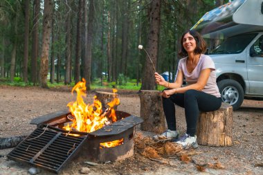 Banff Ulusal Parkı 'ndaki kamp alanında kamp ateşinde şekerleme kızartan kadın turist, karavanının yanında, doğanın huzur ve sessizliğinin tadını çıkarıyor.