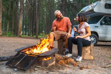 Kamp arabalarının yakınındaki bir kamp ateşinde lokum kızartan turistler, Kanada kayalıklarında, Banff Ulusal Parkı 'nın ormanında rahatlatıcı bir akşamın tadını çıkarıyorlar.