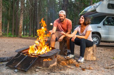 Turistler kamp arabalarının yanındaki sıcak bir kamp ateşinde marşmelov kızartıyorlar. Banff Ulusal Parkı 'nın nefes kesici güzelliği ve görkemli Kanadalı kayalıklarla çevrili.