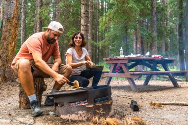 Turistler öğle yemeği için Banff Ulusal Parkı 'ndaki bir kamp alanında ızgara yapıyor. Kanada kayalıkları, Alberta, arka planda piknik masası var.