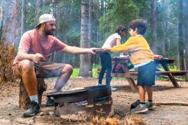 Babam kamp alanında çocuklara yemek servisi yapıyor Banff Ulusal Parkı 'nda kamp ateşinde yemek pişiriyor Kanadalı kayalar, Alberta, yaz tatilinde açık hava yaşam tarzının tadını çıkarıyor.