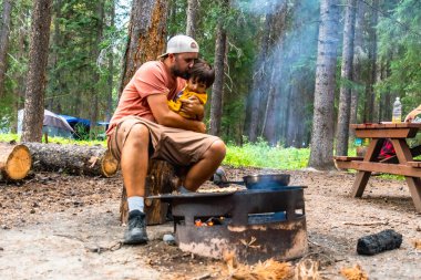 Baba, Banff Ulusal Parkı 'ndaki kamp gezisi sırasında kamp ateşinde kahvaltı yaparken oğluna şefkat gösteriyor. Kanada kayalıkları, Alberta.
