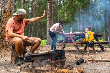 Banff Milli Parkı 'nın nefes kesici manzarasında aile kampı ve ızgara yaparken bir baba el sallıyor, bir anne yemek hazırlıyor ve bir çocuk kamp ateşinin yanında yürüyor.