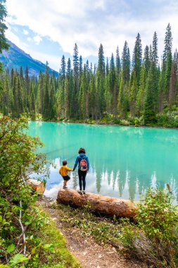 Turistler, Banff Ulusal Parkı, Alberta, Kanada 'daki verimli çam ormanı ve görkemli dağlarla çevrili zümrüt gölünün turkuaz suyunun nefes kesici manzarasının keyfini çıkarıyorlar.