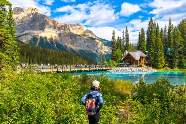 Sırt çantalı kadın turist zümrüt gölünün nefes kesici turkuaz sularına hayran kalıyor. Muhteşem Kanada kayalıklarında ikonik ahşap dağ evi ve köprüsü bulunuyor.