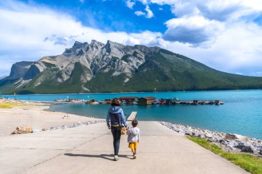 Anne ve çocuk, arka planda kayıklar ve dağlarla Minnewanka Gölü 'ne doğru el ele yürüyorlar. Banff Ulusal Parkı' nda güneşli bir günün tadını çıkarıyorlar.