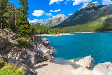Minnewanka gölünün nefes kesici turkuaz suları parlak mavi gökyüzünün altındaki görkemli kayalık dağları yansıtıyor Banff Ulusal Parkı, Alberta, Kanada 'da büyüleyici bir yaz manzarası yaratıyor.