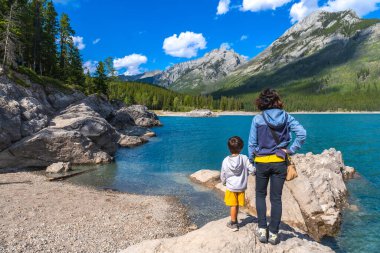 Anne ve oğul, güneşli bir günde, görkemli Kanadalı kayalıklarla çevrili Minnewanka Gölü 'nün büyüleyici turkuaz sularına hayran kalarak bir kayanın üzerinde duruyorlar.