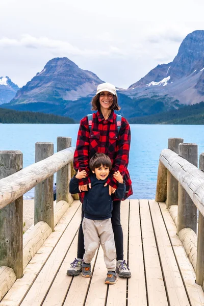 Gülümseyen anne ve oğlu Banff Ulusal Parkı 'ndaki turkuaz yay gölünün ve etrafındaki dağların büyüleyici manzarasını kucaklayarak doğadaki güzel bir aile anını yakalıyorlar.