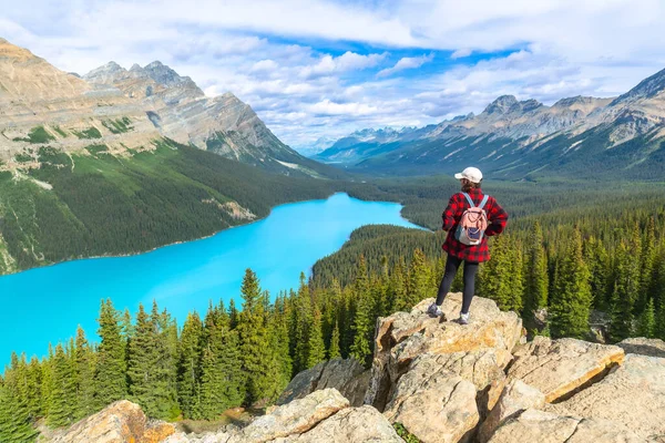 Kayalık bir uçurumda dikilen kadın yürüyüşçü güneşli bir yaz gününde Turkuaz Peyton Gölü 'nün ve Banff Ulusal Parkı' ndaki dağların manzarasının keyfini çıkarıyor.