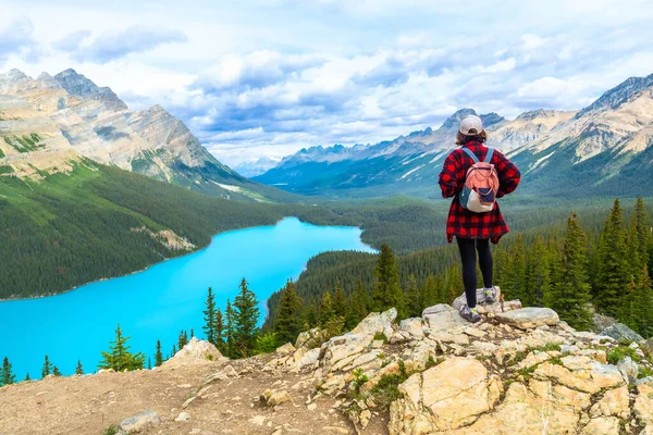 Kayalık bir uçurumda duran kadın yürüyüşçü, bulutlu bir günde Peyton Gölü 'nün turkuaz sularına ve Banff Ulusal Parkı' ndaki dağlara bakıyor.