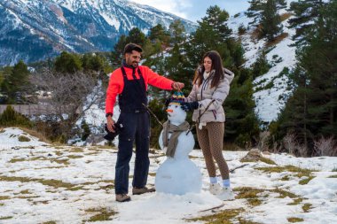 Genç çift, karlı bir dağ manzarasında birlikte bir kardan adam inşa ederek kış aktivitelerini kutluyor ve doğada paylaşılan tatil deneyiminin tadını çıkarıyor.