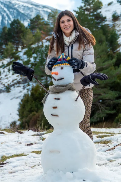 Genç bir kadın şapkası, atkısı, havuç burnu ve ağaç dalları olan bir kardan adam yaratırken gülümsüyor, karlı bir dağ manzarasında kış tatillerini kutluyor.