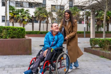 Kadın bakıcı tekerlekli sandalyedeki mutlu bir yetişkini itiyor, şehir parkında eğleniyor, arkadaşlık, destek ve kapsayıcı bir yaşam sergiliyor.