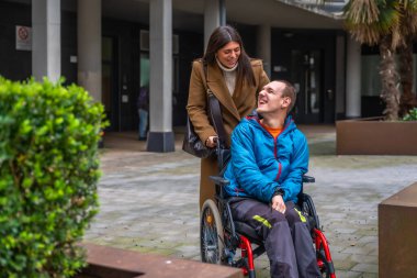 Engelli bir kadın ve erkek, onu tekerlekli sandalyeyle asfalt bir alanda iterken mutlu bir şekilde etkileşime geçiyorlar. Destek, bakım ve aktif bir yaşam tarzını simgeliyorlar.