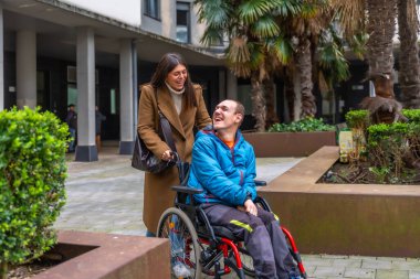 Genç bir kadın mutlu bir şekilde tekerlekli sandalyedeki orta yaşlı bir adamı kaldırımlı bir yola itiyor. Hem gülümsüyor hem de neşeli bir bakım ve destek anı yaşıyor.