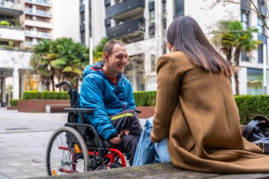 Tekerlekli sandalye kullanan gülümseyen bir adam, şehir ortamında arkadaşlığın, dahil olmanın ve sosyal bağın temalarını yansıtan neşeli bir açık hava sohbetine girişir.