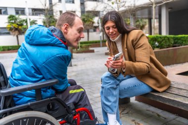 Tekerlekli sandalyede engelli genç bir adam cep telefonuyla içeriğini paylaşan genç bir kadınla etkileşime geçiyor, dış mekanda kapsama, bakım ve dostluğu teşvik ediyor.