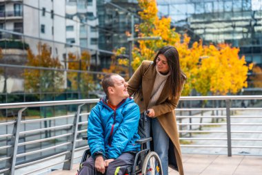 Engelli kadın ve erkek, dışarıda geçen bir sonbahar gününün tadını çıkarıyorlar. Tekerlekli sandalyesini modern şehir ortamında iterken mutlu bir anı paylaşıyorlar. Destek ve arkadaşlığı temsil ediyorlar.