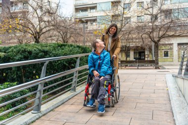 Genç bir kadın tekerlekli sandalyede bir çocuğu itiyor, birlikte gülüyor ve yeşil çalılar ve tırabzan ile ulaşılabilir bir şehir yolunda yürüyor, destek ve kapsama alanının altını çiziyor.