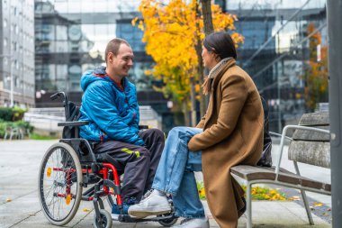 Açık havada neşeli bir sohbete girişen iki yetişkin, sonbahar günü parkta bankta oturan bir kadınla konuşurken tekerlekli sandalyede gülümseyen bir adam.