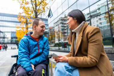 Akıllı telefon tutan bir kadın, tekerlekli sandalyede oturan engelli bir adama bakıyor. Erişilebilirliği, dahil edilmeyi ve dostça bir sohbeti temsil ediyor.