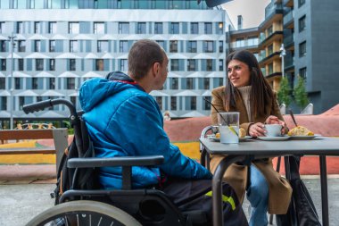 Tekerlekli sandalyedeki genç kadın ve adam açık bir kafede sıcak bir sohbet ve kahve paylaşıyor.