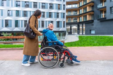 Bakıcı, tekerlekli sandalyedeki gülümseyen bir adamı ulaşılabilir bir şehir parkına itiyor. Kapsamlı, aktif bir şehir gezisi sırasında kahkahalarını ve desteğini paylaşıyor.