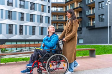 Kadın bakıcı, şehir kaldırımında tekerlekli sandalyeli orta yaşlı bir adamı itiyor, destek ve bağımsızlık sunuyor; sonbahar kentsel sahnesi hareketlilik, empati ve günlük yaşamı gösteriyor