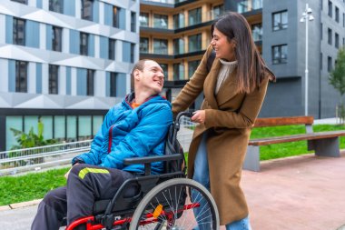 Gülümseyen genç kadın, tekerlekli sandalyedeki adamı şehir caddesi boyunca iterek şehir binaları ve park manzaraları arasında neşeli, destekleyici bir arkadaşlık ve bakım anını paylaşıyor.