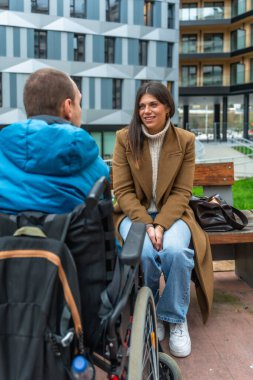 Genç bir kadın park bankında oturmuş tekerlekli sandalyedeki bir adamla konuşuyor, sıcak, kapsayıcı bir anı paylaşıyor destek, ulaşılabilirlik ve kentsel dostluğu ön plana çıkarıyor.
