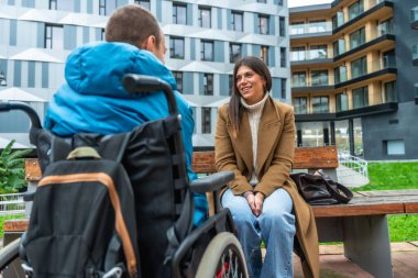 Kadın şehir merkezinde bir bankta oturuyor, gülümsüyor ve tekerlekli sandalye kullanan bir adamla sohbet ediyor, destek ve kapsama temalarını temsil ediyor.