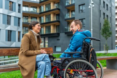Genç bir kadın bankta oturuyor, tekerlekli sandalyedeki orta yaşlı bir adamla neşeli bir konuşma yapıyor, hem gülüyor hem de şehir parkında birbirine bakıyor.