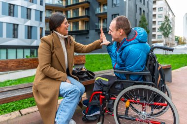 Tekerlekli sandalyede engelli genç bir kadın ve yetişkin bir adam beşlik çakıyor, gülümsüyor ve bir şehir parkında destek veriyor.