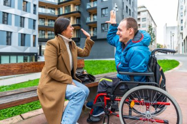 Kadın bankta oturuyor ve erkek tekerlekli sandalyede gülümsüyor, birbirine beşlik çakıyor, ve şehir parkında mutlu bir anı paylaşıyor, destek, dostluk ve kapsama gösteriyor.