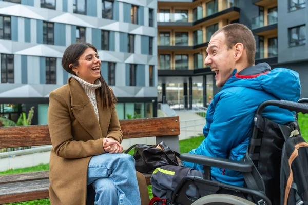 Genç bir kadın bankta oturmuş tekerlekli sandalyedeki orta yaşlı bir adamla keyifli bir sohbetin tadını çıkarıyor.