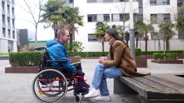 Tekerlekli sandalyede sakat genç bir adam, modern şehir parkında bir bankta oturan bir bayan arkadaşıyla keyifli ve olumlu bir konuşma yapıyor.