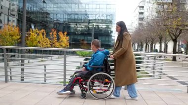 Tekerlekli sandalyedeki genç adam ve kadın bakıcısı bir sonbahar günü arka planda kentsel mimariyle birlikte modern bir şehir caddesinde geziniyorlar.
