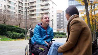 Tekerlekli sandalyedeki genç adam, şehir parkındaki bir kadın bakıcı ya da arkadaşıyla canlı ve mutlu bir konuşma yapıyor, destekleyici ilişkiler ve pozitif sosyal etkileşim sergiliyor.