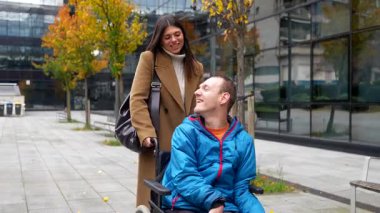 Tekerlekli sandalyedeki mutlu sakat adam bakıcısıyla şehirde keyifli bir yürüyüş yapıyor. Engelli hastasını sonbaharda dışarı iterken konuşan ve gülümseyen bir asistan.