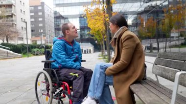 Tekerlekli sandalyede oturan engelli genç bir adam, şehir bankında bir bayan arkadaşıyla sohbet ediyor, şehir ortamında bir bağlantı ve destek anı paylaşıyor.