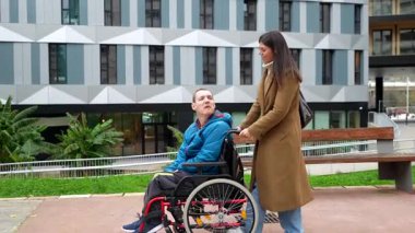 Genç bir bakıcı kadın tekerlekli sandalyede fiziksel engelli bir adamı şehir parkına itiyor. Destek, ulaşılabilirlik ve olumlu ilişkileri temsil ediyor.