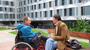 Fiziksel engelli genç bir adam tekerlekli sandalyede oturuyor ve kadın bakıcısıyla açık hava terapisi yapıyor, modern bir binanın önündeki bankta konuşuyor ve gülümsüyor.